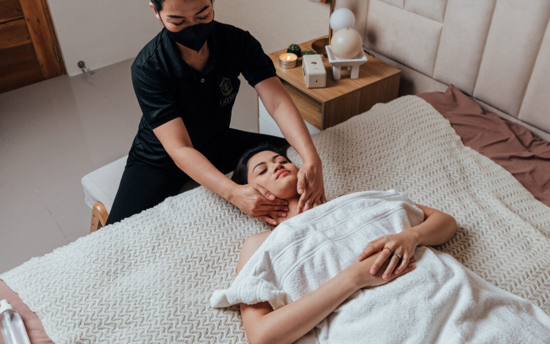 A person receives a relaxing face massage from a therapist wearing a black mask. They lie on a bed covered in a white towel, surrounded by calming decor.