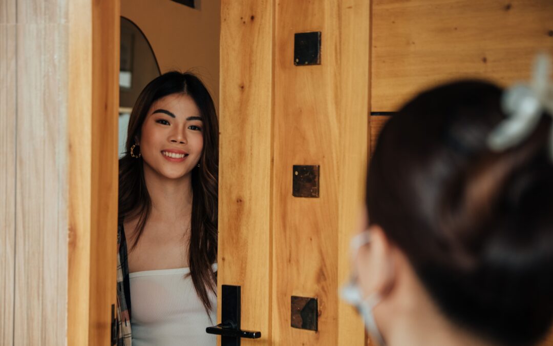 A woman with long hair and hoop earrings smiles warmly at the open wooden door. Another person's head, blurred in the foreground, faces her.